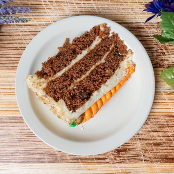 A slice of carrot cake with white frosting and an orange carrot decoration on a white plate.