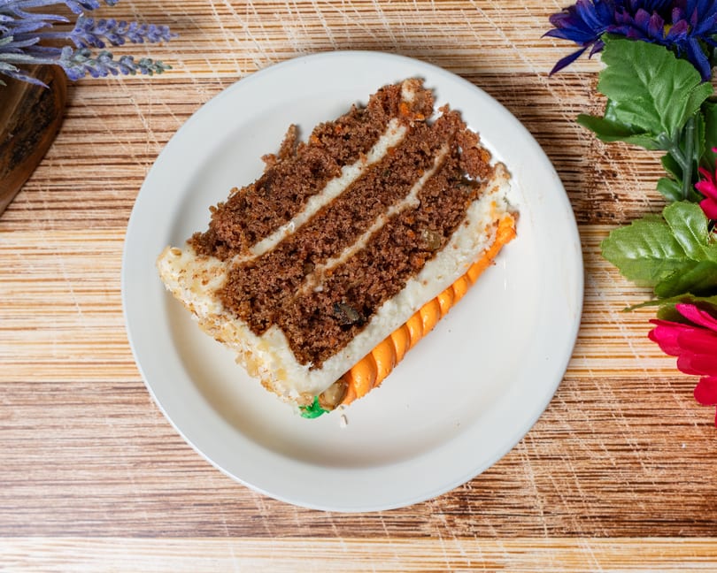 A slice of carrot cake with white frosting and an orange carrot decoration on a white plate.