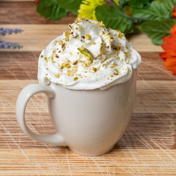 A white mug filled with whipped cream and topped with chopped pistachios on a wooden surface.