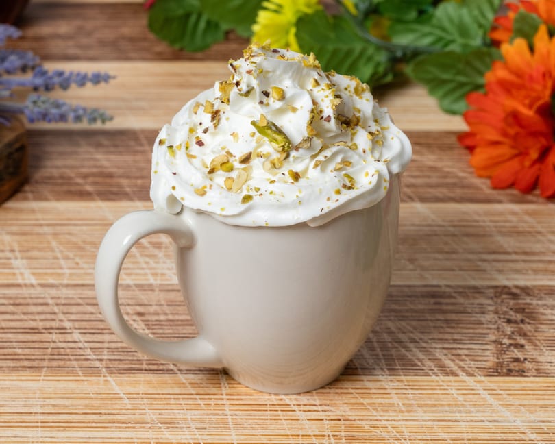 A white mug filled with whipped cream and topped with chopped pistachios on a wooden surface.