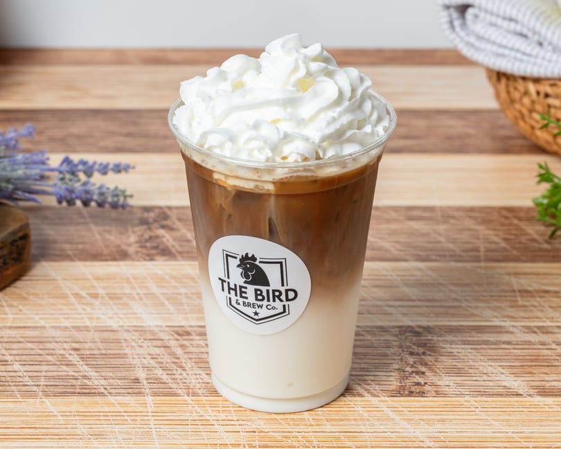 Iced coffee with whipped cream and "The Bird & Brew Co." logo on a wooden surface.