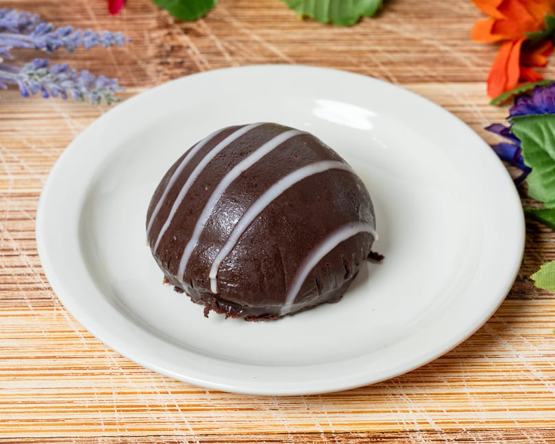 A dark chocolate dessert with white icing stripes on a white plate, on a wooden surface.