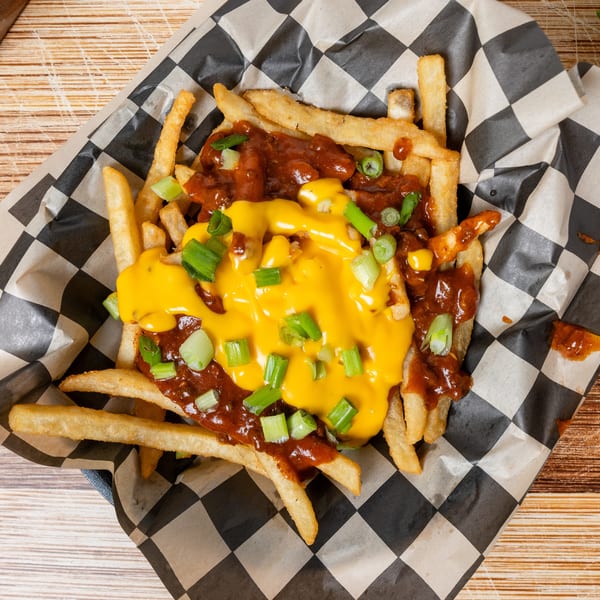 Chili cheese fries with chopped green onions in a checkered paper-lined basket on a wooden table.
