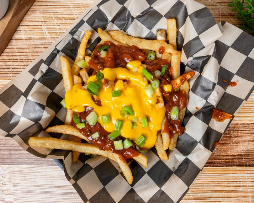 Chili cheese fries with chopped green onions in a checkered paper-lined basket on a wooden table.